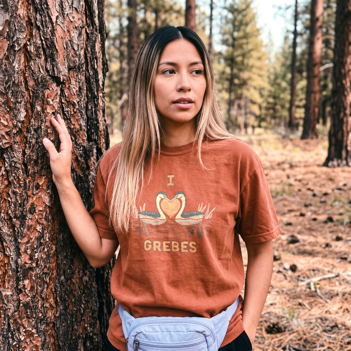 A woman standing outdoors against a large tree trunk in a forest, wearing a rust-orange graphic T-shirt with a grebe design and the word 'I LOVE GREBES' on the front, plus a light blue fanny pack.