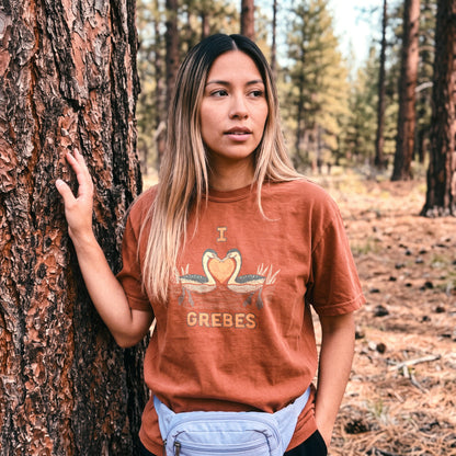 A woman standing outdoors against a large tree trunk in a forest, wearing a rust-orange graphic T-shirt with a grebe design and the word 'I LOVE GREBES' on the front, plus a light blue fanny pack.